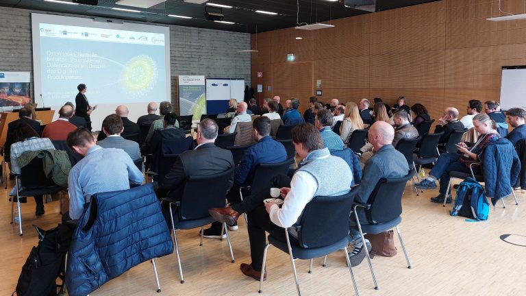 In einem modernen Konferenzraum sitzen rund 40 Personen in Reihen und verfolgen einen Vortrag. Vorne steht eine präsentierende Person neben einer großen Leinwand mit der Folie „Daten teilen, Kontrolle behalten: Potenziale von Datenräumen am Beispiel des Digitalen Produktpasses“. Rechts an der Wand befinden sich Holzpaneele, links ist eine Betonwand zu sehen.