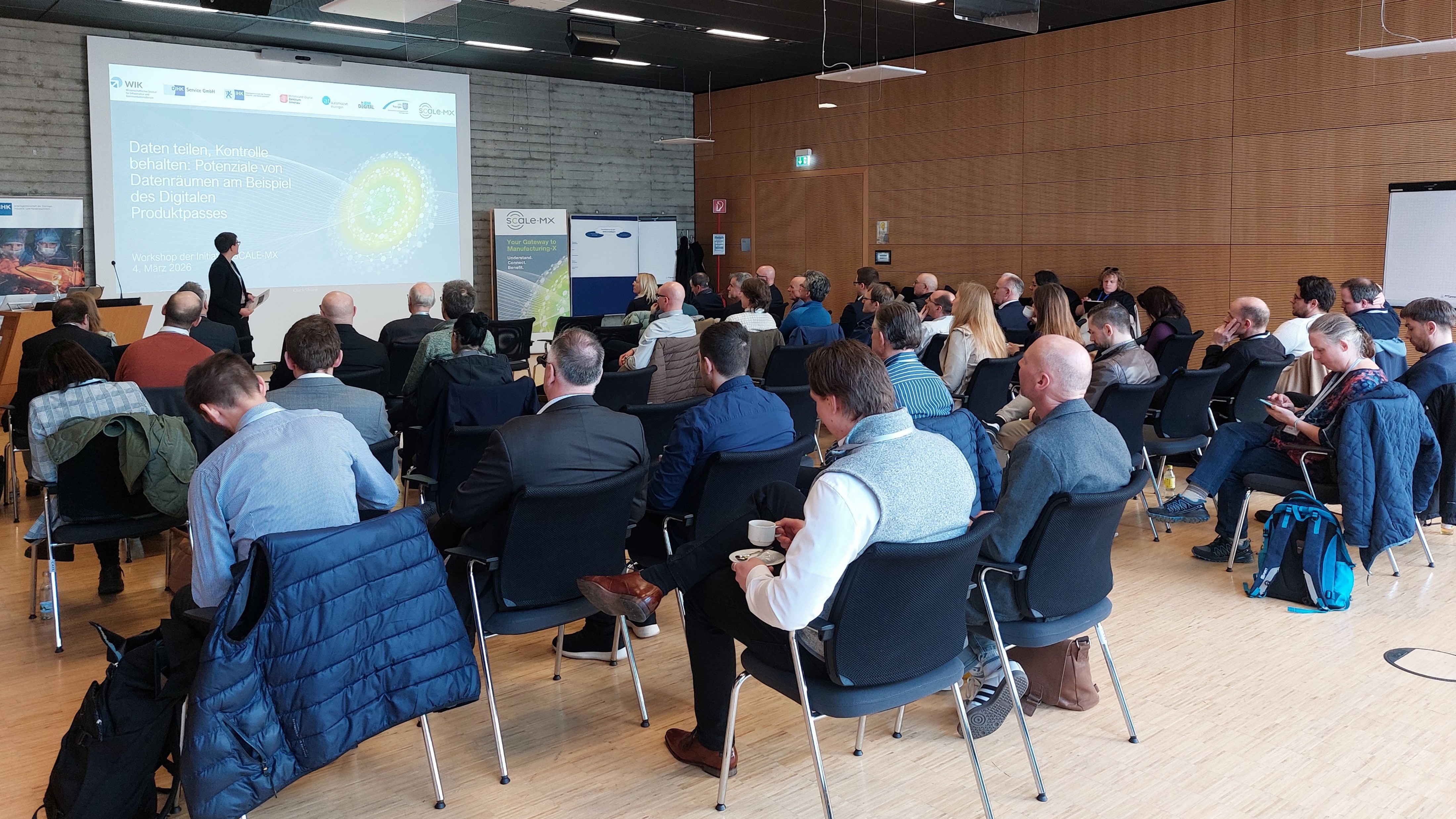 In einem modernen Konferenzraum sitzen rund 40 Personen in Reihen und verfolgen einen Vortrag. Vorne steht eine präsentierende Person neben einer großen Leinwand mit der Folie „Daten teilen, Kontrolle behalten: Potenziale von Datenräumen am Beispiel des Digitalen Produktpasses“. Rechts an der Wand befinden sich Holzpaneele, links ist eine Betonwand zu sehen.