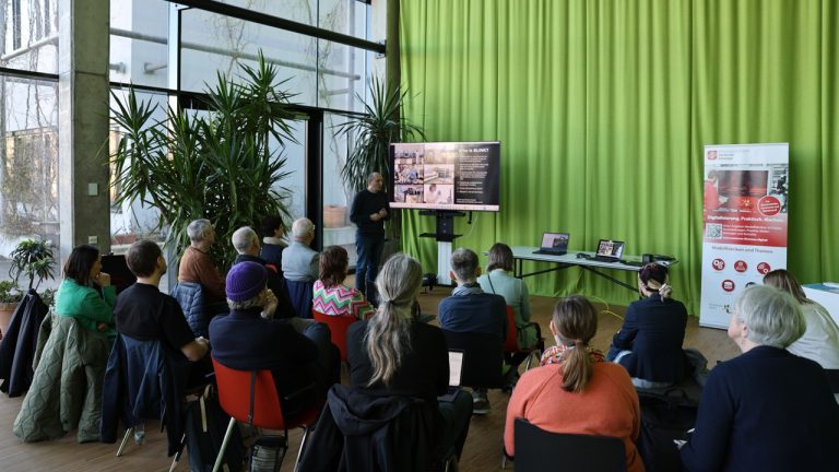 Vortrag Entdeckertour in Jena In einem hellen Veranstaltungsraum mit großer Fensterfront und grünen Vorhängen sitzen mehrere Personen auf Stühlen und hören einem Vortrag zu. Vorne steht ein Referent neben einem Bildschirm mit Präsentationsfolie; rechts daneben stehen ein Roll-up-Banner und ein Tisch mit Laptops. Große Zimmerpflanzen prägen den Raum.