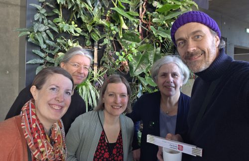 Fünf Personen lächeln in die Kamera und stehen vor einer begrünten Pflanzenwand in einem Innenraum. Eine Person hält eine Tasse und mehrere Broschüren in der Hand. Die Gruppe wirkt locker und steht eng beieinander für ein gemeinsames Foto.
