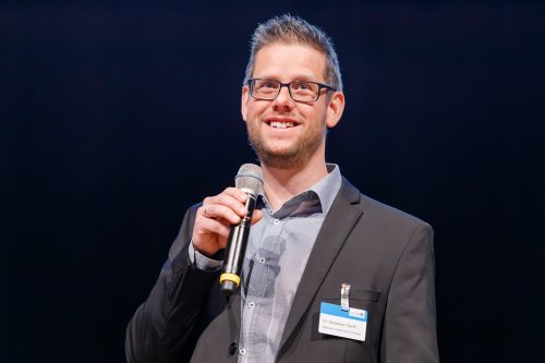 Dr. Sebastian Gerth steht mit einem Mikro in de Hand auf der Bühne