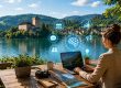 Eine Frau arbeitet an einem Laptop auf einer Terrasse am See, während im Hintergrund eine schlichte Burg vor bewaldeten Mittelgebirgshängen zu sehen ist; digitale Symbole visualisieren den Einsatz von KI im Tourismus.