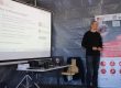 KI Trainer Wolfram Kattanek hält einen Vortrag neben einer Präsentationsleinwand mit Informationen zu „Mittelstand-Digital“. Rechts daneben steht ein Roll-up-Banner des Mittelstand-Digital Zentrums Ilmenau zum Thema intelligente Sensorsysteme. Vor dem Vortragenden befinden sich ein Tisch mit Laptop und Lautsprechern sowie mehrere Stühle in einem provisorisch aufgebauten Veranstaltungsraum.