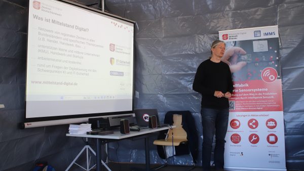 KI Trainer Wolfram Kattanek hält einen Vortrag neben einer Präsentationsleinwand mit Informationen zu „Mittelstand-Digital“. Rechts daneben steht ein Roll-up-Banner des Mittelstand-Digital Zentrums Ilmenau zum Thema intelligente Sensorsysteme. Vor dem Vortragenden befinden sich ein Tisch mit Laptop und Lautsprechern sowie mehrere Stühle in einem provisorisch aufgebauten Veranstaltungsraum.