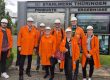 Gruppenfoto von acht Personen vor dem Eingangsschild des Stahlwerks Thüringen. Die Teilnehmenden tragen orange Schutzmäntel und weiße Helme, im Hintergrund sind Infotafeln mit Produkten und Ergebnissen des Werks zu sehen.