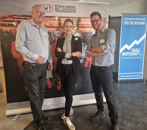 Drei Personen stehen vor einem Banner des Stahlwerks Thüringen und einem Roll-up des Unternehmernetzwerks Saale Wirtschaft. Die Szene wirkt wie ein Gruppenfoto im Rahmen einer Netzwerk- oder Informationsveranstaltung.