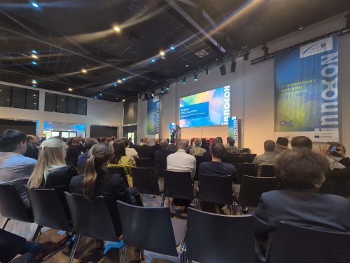Blick in einen gut besuchten Konferenzsaal während der innoCON, der Innovationskonferenz in Thüringen. Auf der Bühne stehen zwei Personen vor einer Präsentationsfolie mit dem Titel „Grußwort“, daneben sind große innoCON-Banner zu sehen.