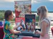 Zwei Personen stehen an einem Informationsstand des Mittelstand-Digital Zentrums Ilmenau und der Ernst-Abbe-Hochschule Jena. Im Mittelpunkt steht ein Demonstrator zum Thema „Alternativ investieren: Kaffee, Kautschuk oder Kupfer – Was sagt die KI?“, im Hintergrund ist ein See zu sehen.