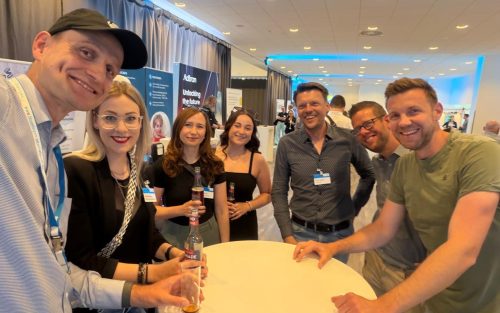 Selfie einer Gruppe von sieben Personen an einem Stehtisch im Ausstellungsbereich einer Konferenz. Im Hintergrund sind Messestände, Roll-ups und weitere Teilnehmende zu sehen. Die Stimmung wirkt locker und freundlich.