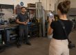 Eine Frau filmt mit ihrem Smartphone einen Handwerker, der vor einer Werkbank in einer gut ausgestatteten Werkstatt steht und in die Kamera spricht.