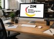 Moderner Computer auf einem Schreibtisch in einer Büroumgebung. Auf dem Bildschirm ist das Logo des Zentralen Innovationsprogramms Mittelstand (ZIM) mit den Schriftzügen „ZIM“ und „Zentrales Innovationsprogramm Mittelstand“ zu sehen. Auf dem Schreibtisch befinden sich Tastatur, Maus, Notizbuch, Stifte, eine Schreibtischlampe sowie Pflanzen im Hintergrund.
