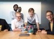 Mehrere Personen sitzen und stehen um einen Besprechungstisch in einem Konferenzraum. Auf dem Tisch stehen geöffnete Laptops mit Aufklebern, Tassen und ein weiteres geschlossenes Notebook. Im Hintergrund ist ein Whiteboard zu sehen. Die Personen sind auf die Besprechung ausgerichtet und befinden sich in einer kollaborativen Arbeitssituation.