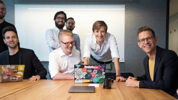 Startup-Meeting Mehrere Personen sitzen und stehen um einen Besprechungstisch in einem Konferenzraum. Auf dem Tisch stehen geöffnete Laptops mit Aufklebern, Tassen und ein weiteres geschlossenes Notebook. Im Hintergrund ist ein Whiteboard zu sehen. Die Personen sind auf die Besprechung ausgerichtet und befinden sich in einer kollaborativen Arbeitssituation.