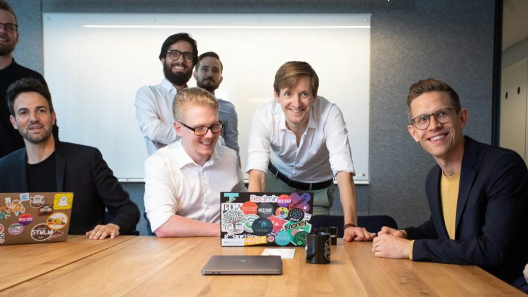 Startup-Meeting Mehrere Personen sitzen und stehen um einen Besprechungstisch in einem Konferenzraum. Auf dem Tisch stehen geöffnete Laptops mit Aufklebern, Tassen und ein weiteres geschlossenes Notebook. Im Hintergrund ist ein Whiteboard zu sehen. Die Personen sind auf die Besprechung ausgerichtet und befinden sich in einer kollaborativen Arbeitssituation.