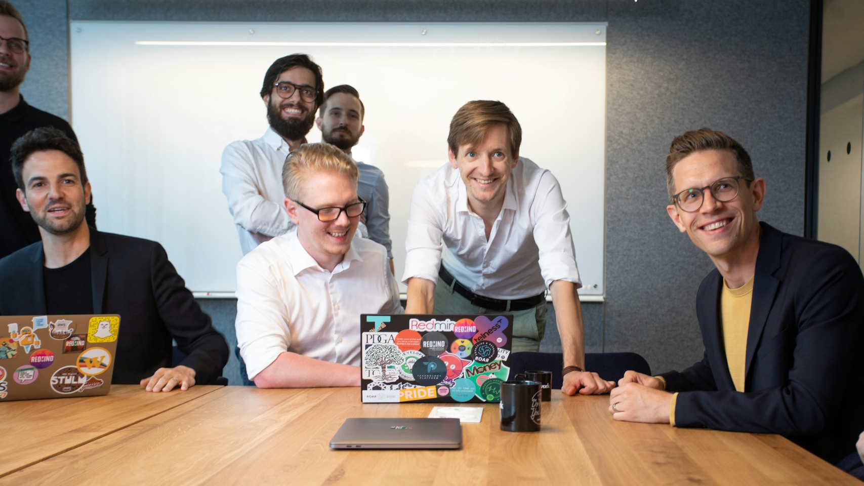 Mehrere Personen sitzen und stehen um einen Besprechungstisch in einem Konferenzraum. Auf dem Tisch stehen geöffnete Laptops mit Aufklebern, Tassen und ein weiteres geschlossenes Notebook. Im Hintergrund ist ein Whiteboard zu sehen. Die Personen sind auf die Besprechung ausgerichtet und befinden sich in einer kollaborativen Arbeitssituation.