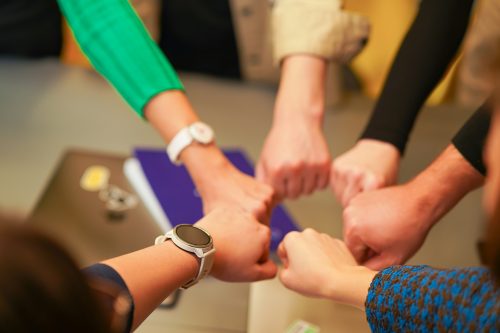 Mehrere Personen strecken ihre Fäuste in die Mitte eines Tisches und berühren sich in einer gemeinsamen Geste. Sichtbar sind Arme mit unterschiedlicher Kleidung sowie eine Armbanduhr. Im Hintergrund liegen ein Notizbuch und weitere Gegenstände auf dem Tisch. Das Bild vermittelt Zusammenarbeit und gemeinsames Handeln.