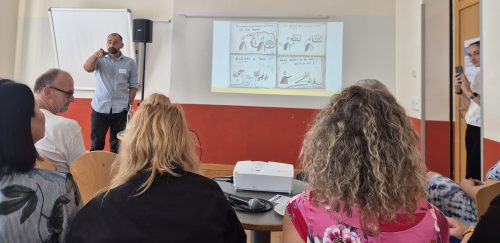 Ein Referent spricht vor einer Gruppe über Kommunikationsprobleme und Konflikte im Team, während eine Präsentation an die Wand projiziert wird.