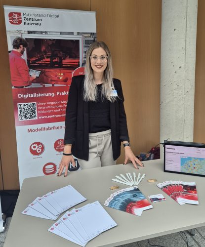 Mitarbeiterin des Mittelstand-Digital Zentrums Ilmenau steht an einem Informationsstand mit Flyern, Broschüren und einem Roll-up zur Digitalisierung.