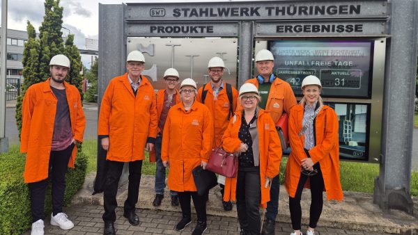 Gruppenfoto von acht Personen vor dem Eingangsschild des Stahlwerks Thüringen. Die Teilnehmenden tragen orange Schutzmäntel und weiße Helme, im Hintergrund sind Infotafeln mit Produkten und Ergebnissen des Werks zu sehen.