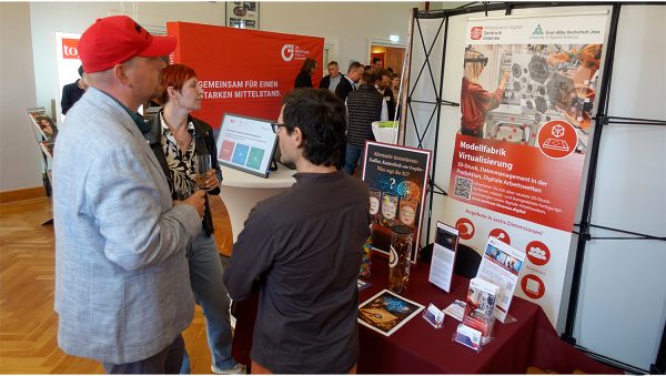 Mehrere Personen stehen an einem Informationsstand des Mittelstand-Digital Zentrums Ilmenau und der Ernst-Abbe-Hochschule Jena. Zu sehen sind Roll-ups zur Modellfabrik Virtualisierung, Informationsmaterialien und ein Demonstrator zum Thema alternatives Investieren mit KI.