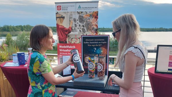 Zwei Personen stehen an einem Informationsstand des Mittelstand-Digital Zentrums Ilmenau und der Ernst-Abbe-Hochschule Jena. Im Mittelpunkt steht ein Demonstrator zum Thema „Alternativ investieren: Kaffee, Kautschuk oder Kupfer – Was sagt die KI?“, im Hintergrund ist ein See zu sehen.