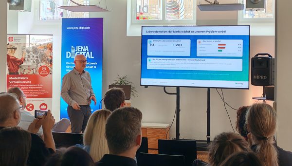 Person steht vor einem großen Bildschirm und hält einen Vortrag in einem Veranstaltungsraum. Auf dem Bildschirm ist eine Präsentationsfolie zum Thema Laborautomation zu sehen. Mehrere Zuhörende sitzen im Publikum. Im Hintergrund stehen Informationsbanner, darunter ein Banner von Jena Digital. Helle Fenster und Veranstaltungstechnik rahmen die Szene ein.